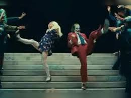 Joaquin Phoenix as Joker and Lady Gaga as Harley Quinn dancing on stairs in 'Joker: Folie à Deux', surrounding by dancers.